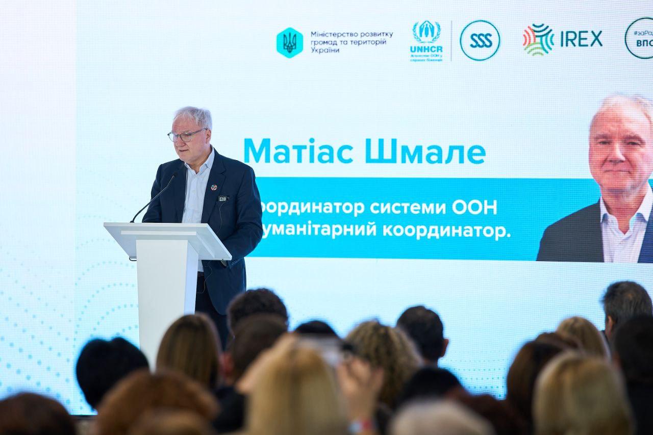 Чоловік у костюмі (Координатор системи ООН) виступає з трибуни перед аудиторією. На великому екрані позаду нього відображено його ім'я, посада та логотипи організацій, включаючи Міністерство розвитку громад та територій України, UNHCR та IREX.