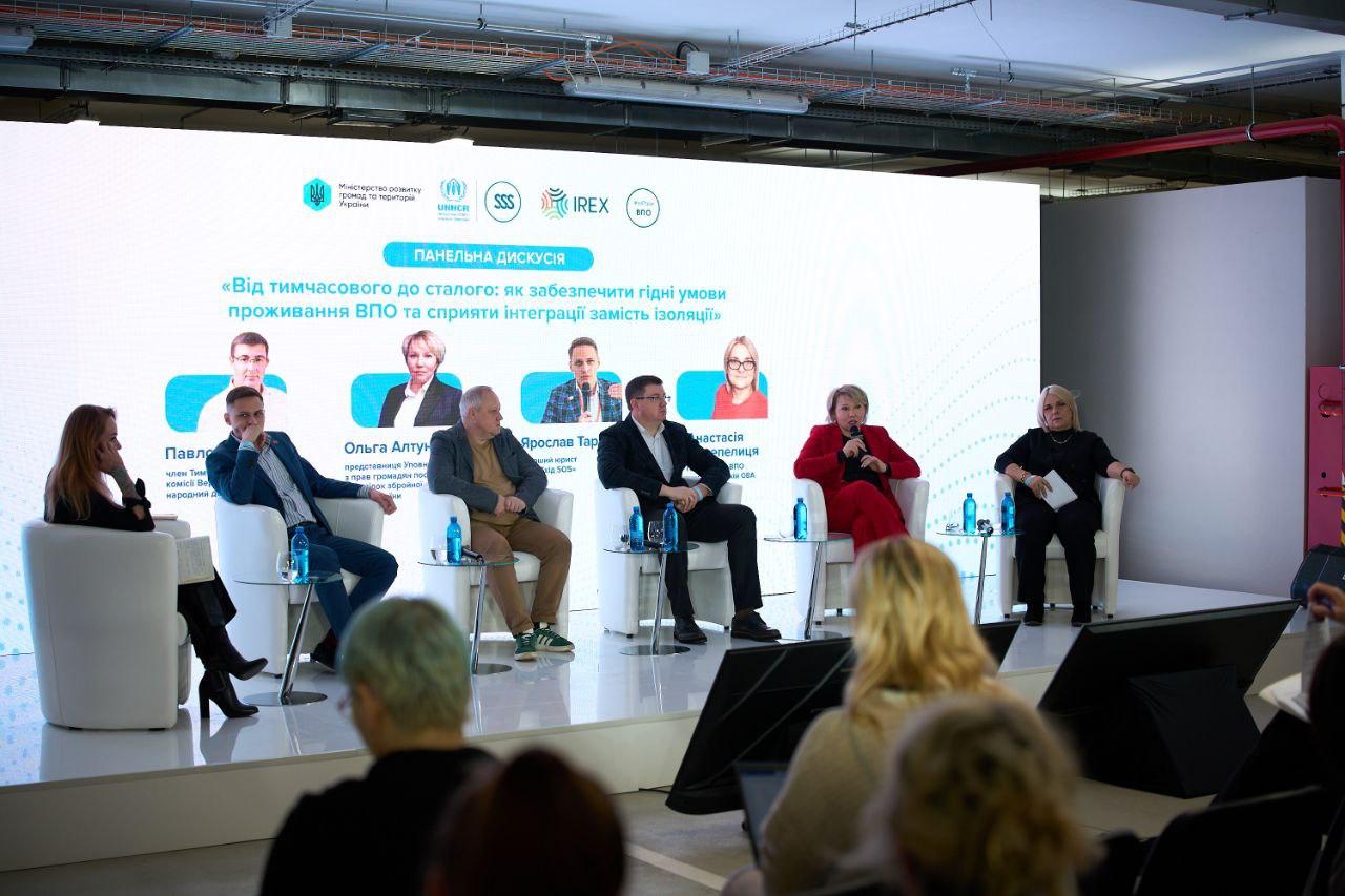 Панельна дискусія на тему: Від тимчасового до сталого: як забезпечити гідні умови проживання ВПО та сприяти інтеграції замість ізоляції. На сцені сидять шість учасників, а на великому екрані позаду них відображені їхні фотографії та імена. Логотипи Міністерства розвитку громад та територій України, UNHCR та IREX видно у верхній частині екрана.