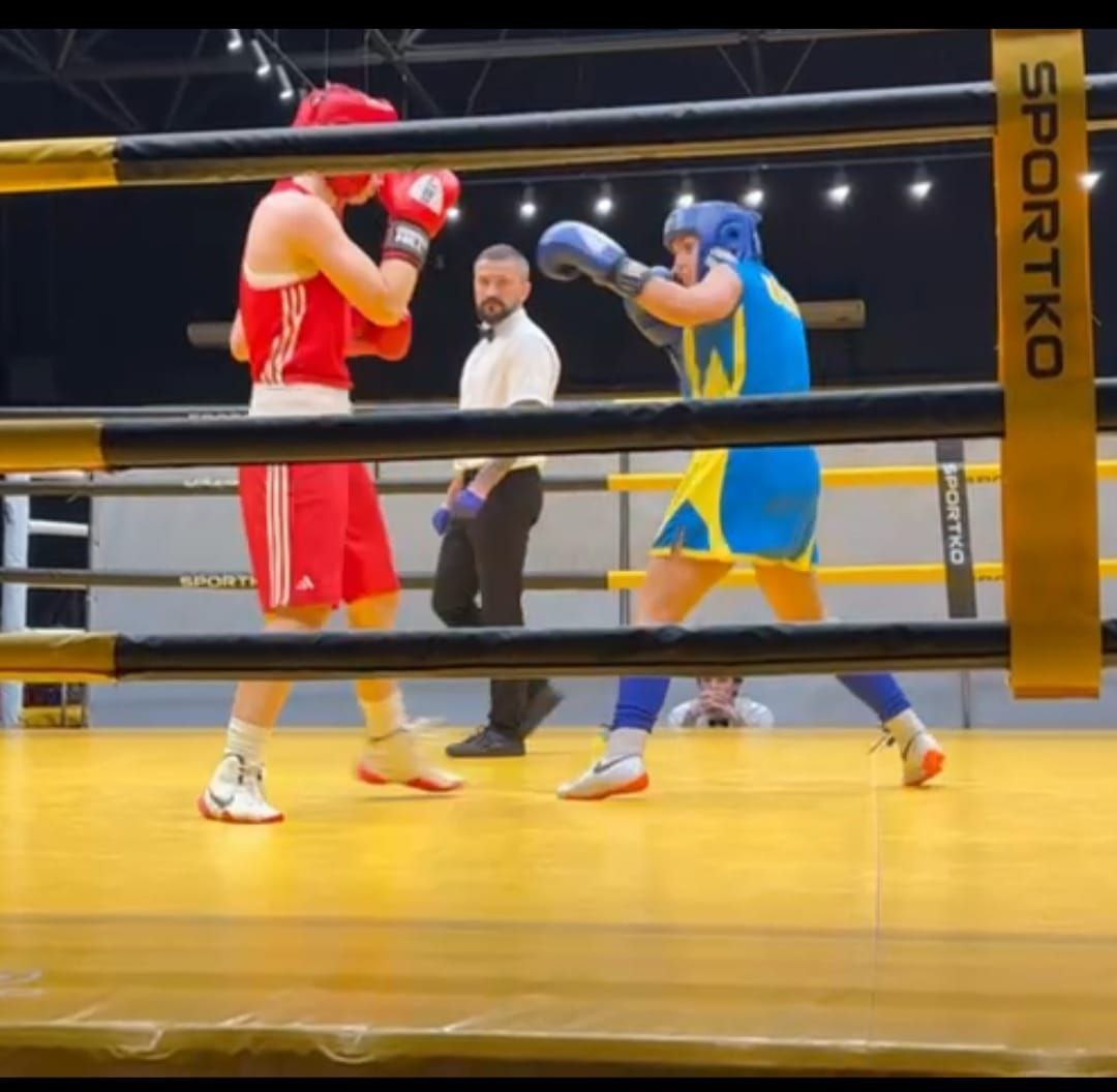 Два боксери у повній екіпіровці (один у червоному, інший у синьо-жовтому) ведуть бій на ринзі. Рефері у білій сорочці стоїть між ними. На кутовій стійці рингу видно напис: SPORTKO.