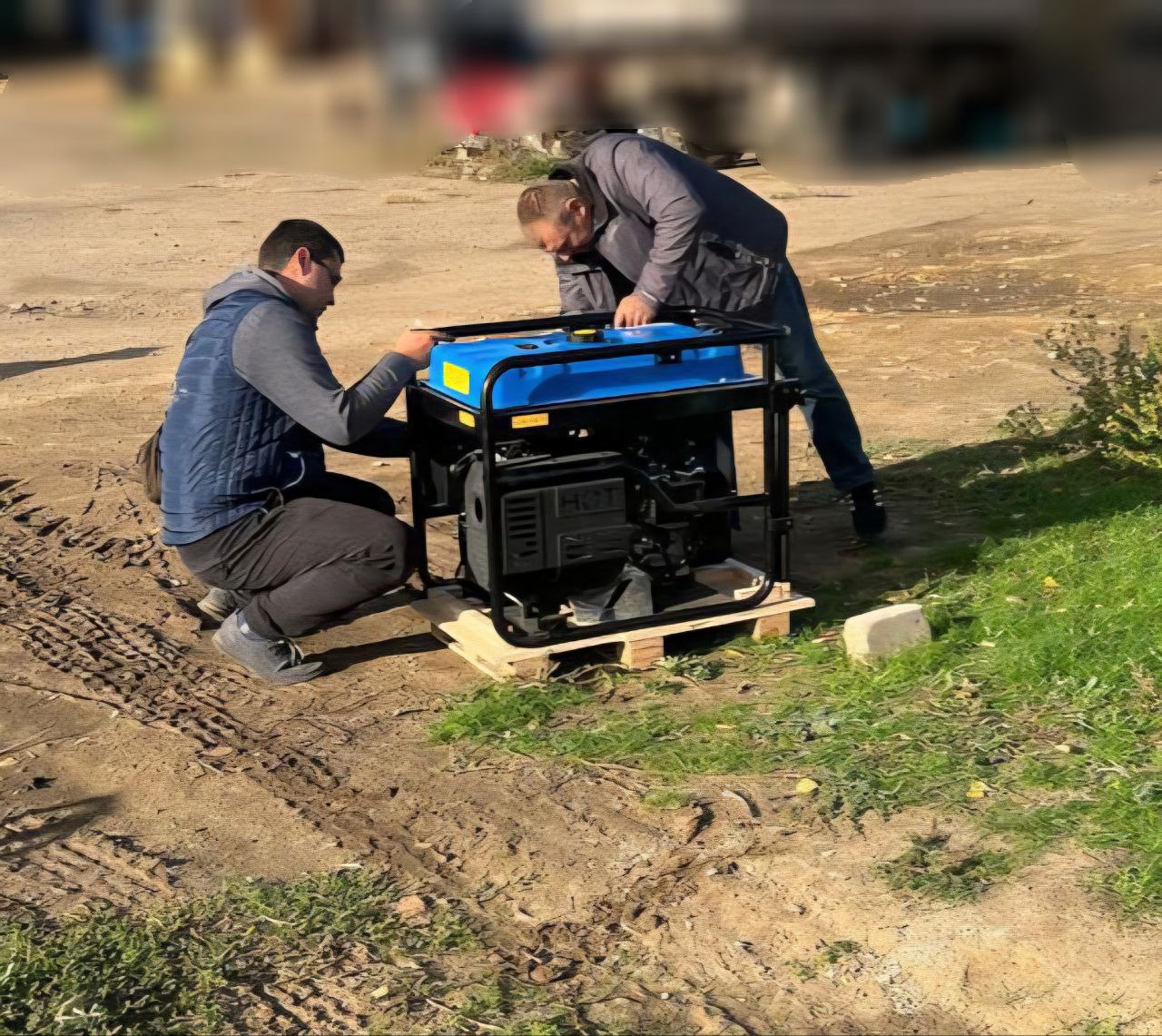 Два чоловіки, один у синьому жилеті, інший у сірій куртці, нахиляються над новим портативним електрогенератором чорно-синього кольору, який стоїть на дерев'яному піддоні на ґрунтовій ділянці.