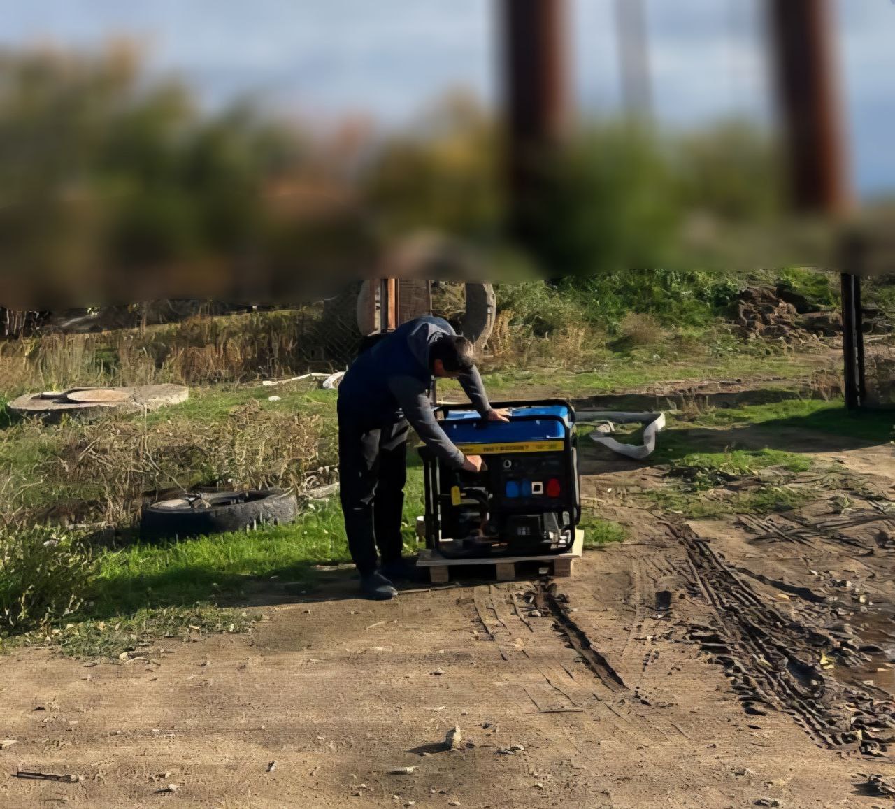 Чоловік у темному одязі нахиляється над синьо-чорним електрогенератором, який стоїть на дерев'яному піддоні на ґрунтовій дорозі, оглядаючи його або підключаючи обладнання.