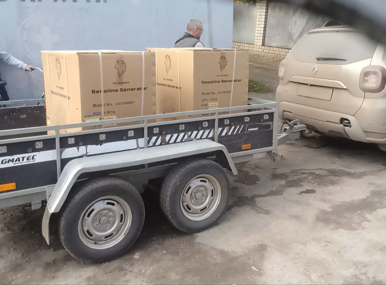 Два великі картонні ящики з написом: Gasoline Generator (Бензиновий генератор) стоять на причепі з двома колесами, причепленому до світлого автомобіля позаду. Чоловік у світлому одязі та окулярах стоїть поруч.