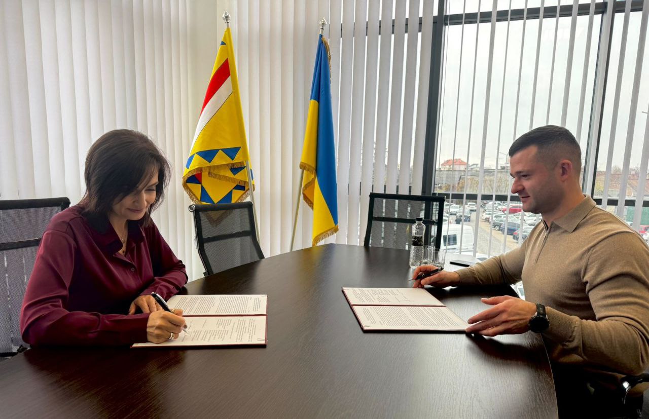 Жінка в бордовій блузці та чоловік у бежевому светрі сидять навпроти один одного за столом і підписують офіційні документи. На задньому плані видно прапори (зокрема, прапор України) та вікно з видом на місто.