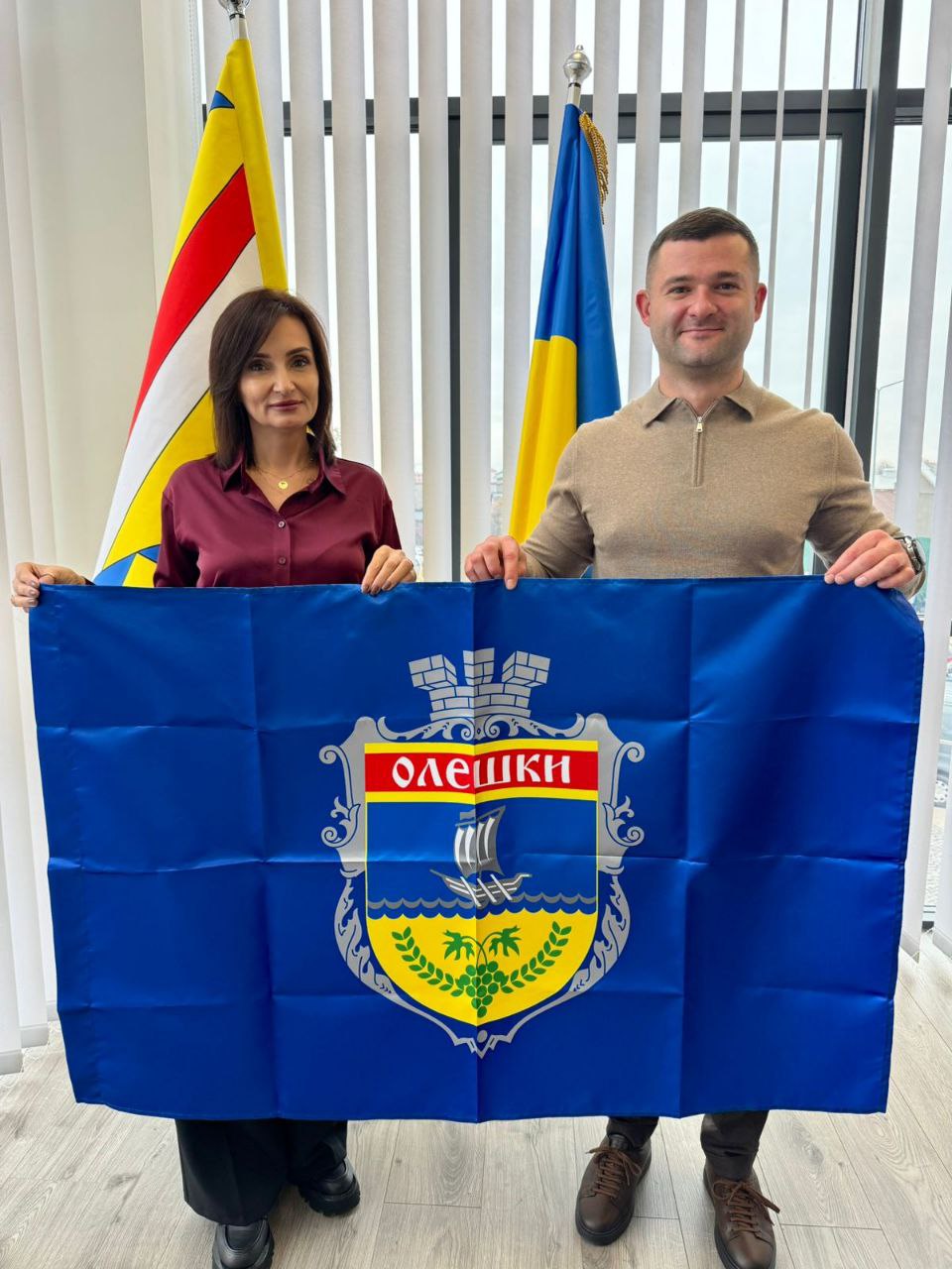 Чоловік у бежевому светрі та жінка в бордовій блузці стоять поруч, демонструючи великий синій прапор із гербом міста Олешки. Позаду них видно державний прапор України та, ймовірно, прапор регіону.