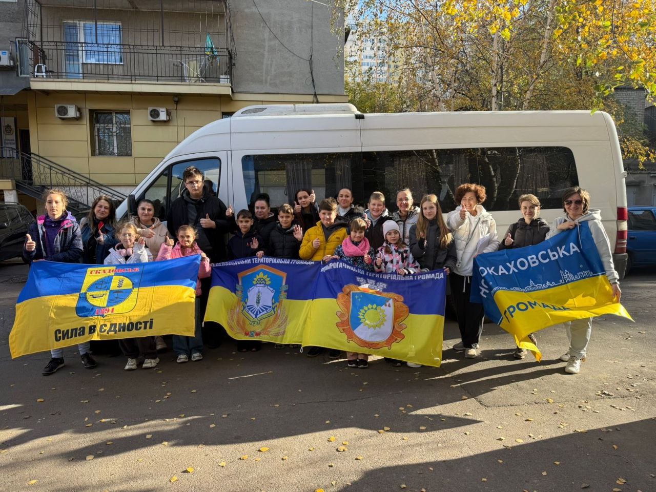 Група дорослих та дітей стоїть перед білим мікроавтобусом, тримаючи великі синьо-жовті прапори з написами, зокрема: Сила в єдності та символікою громад. Багато людей показують великі пальці вгору (жест клас) і посміхаються.