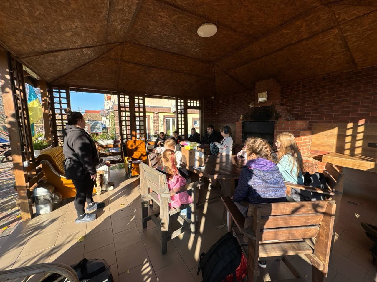 Група дітей та дорослих сидить навколо великого дерев'яного столу в альтанці з дерев'яним дахом та цегляним мангалом. Жінка в чорному одязі стоїть ліворуч, звертаючись до присутніх. Сонячне світло проникає в альтанку з вулиці.