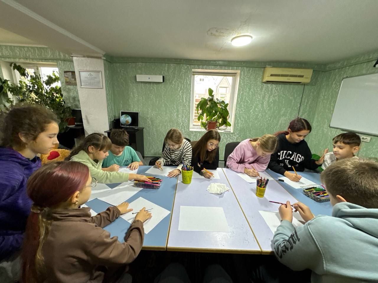 Група з дев'яти дітей та підлітків сидить навколо прямокутного столу та малює на чистих аркушах паперу, використовуючи олівці та фломастери. На столі видно склянки з канцелярським приладдям.