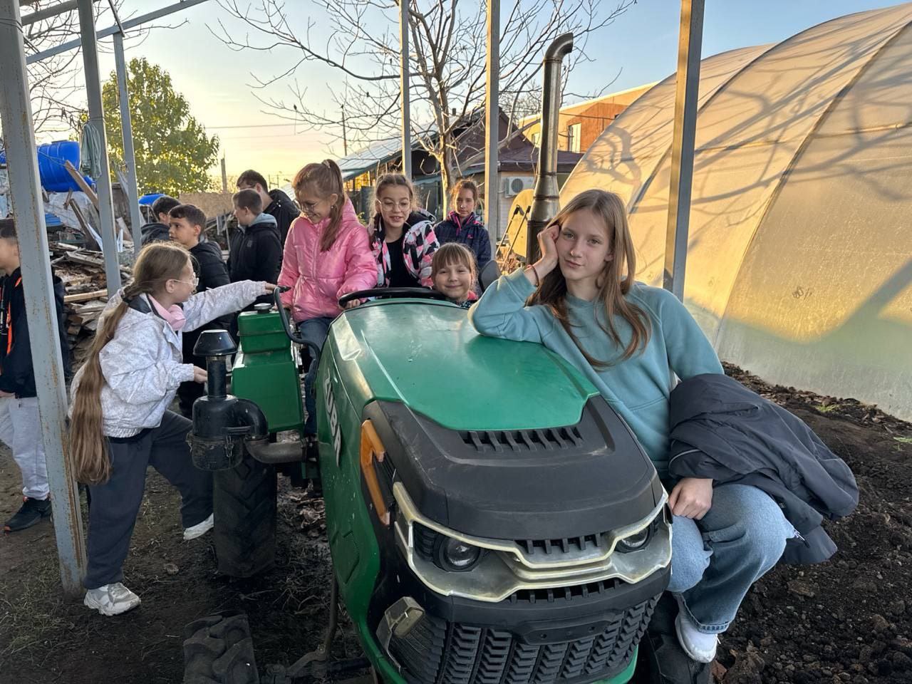 Група усміхнених дітей сидить на зеленому невеликому тракторі або газонокосарці. Дівчинка у світло-блакитній кофті сидить спереду, притуливши руку до обличчя, а інші діти розташовані за нею. На задньому плані видно теплицю та сільськогосподарські угіддя.