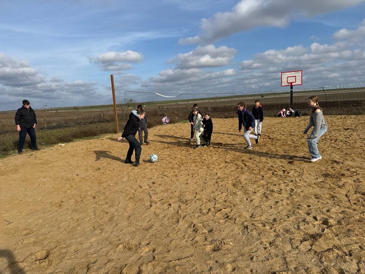 Група дітей та підлітків грає у футбол на піщаному майданчику просто неба під хмарним небом. На задньому плані видно волейбольну сітку та баскетбольний щит.
