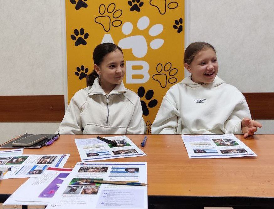 Дві дівчинки-підлітки у світлих кофтах сидять за столом, на якому розкладені навчальні матеріали. Вони дивляться праворуч і посміхаються. На задньому плані — жовтий банер із великими літерами: АВ та відбитками лап.