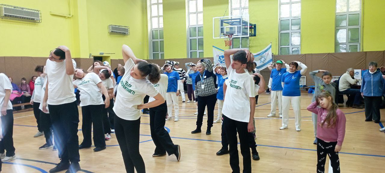 Група людей різного віку, більшість з яких у білих футболках та темних штанах, виконує фізичні вправи (розтяжку) у великому спортивному залі з жовтими стінами та баскетбольними кільцями. На задньому плані видно прапор Ізраїлю.