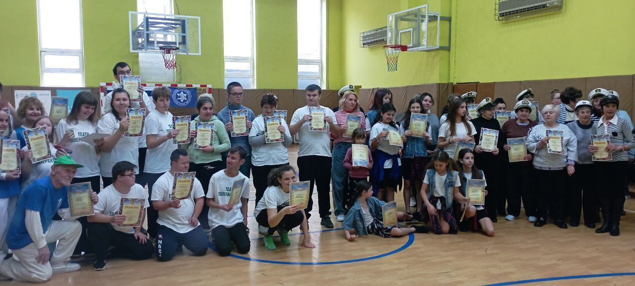 Велика група дорослих та дітей стоїть і сидить на підлозі у спортивному залі, тримаючи в руках сертифікати або подяки. Деякі учасники одягнені у тематичні головні убори (можливо, моряків).