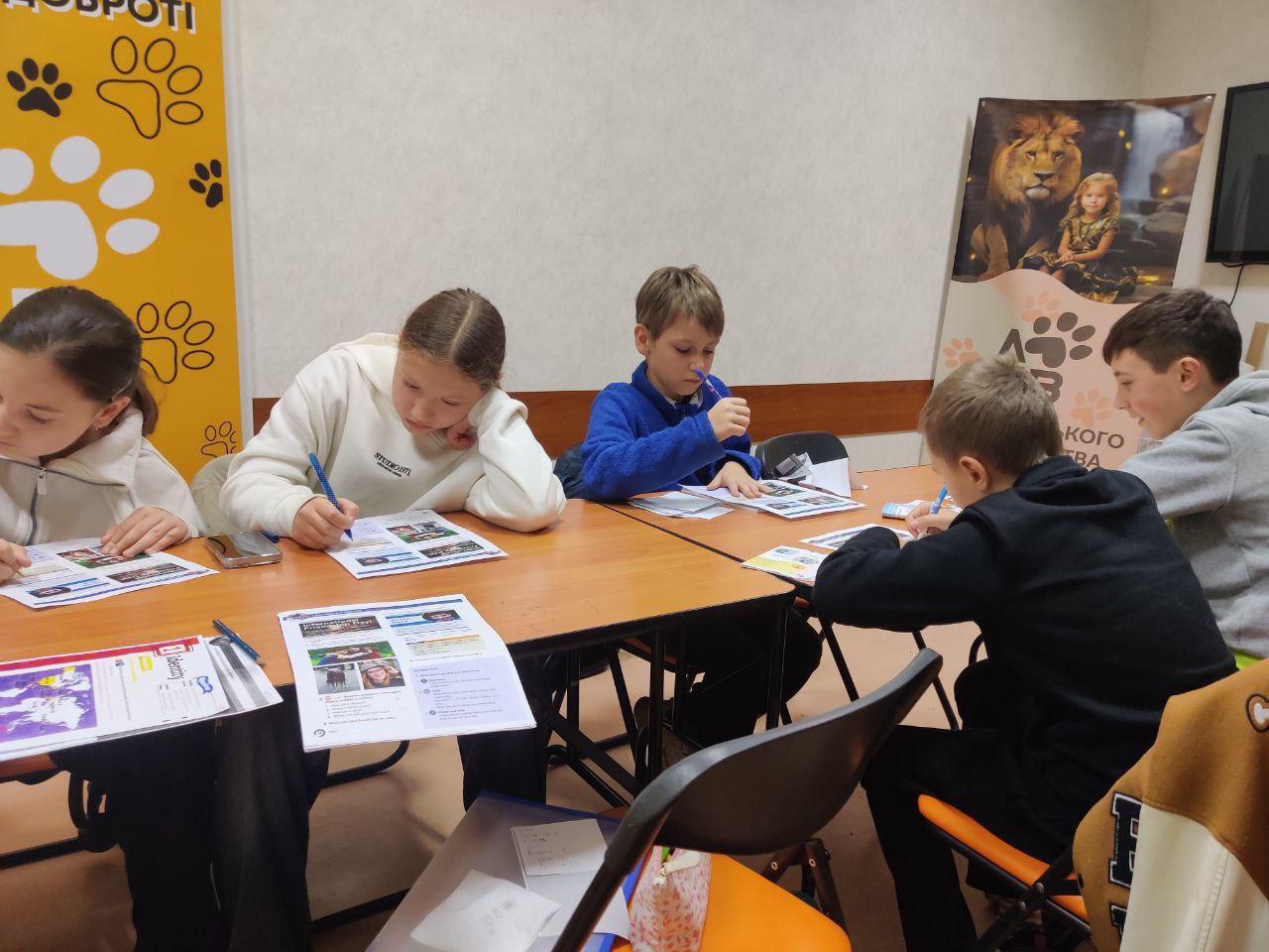 П'ятеро дітей та підлітків сидять за столом і зосереджено працюють із навчальними матеріалами та пишуть у своїх зошитах чи на аркушах. На стіні позаду них видно плакат із зображенням лева, дитини та слідами тварин.