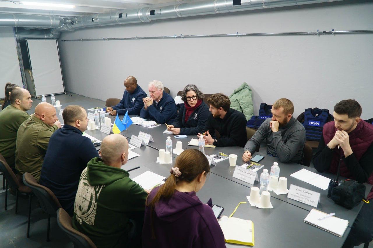 Загальний план офіційної зустрічі у великому світлому залі. Учасники з обох сторін сидять за довгим столом, обговорюючи питання та роблячи нотатки. На столі встановлено прапорці України та ООН.