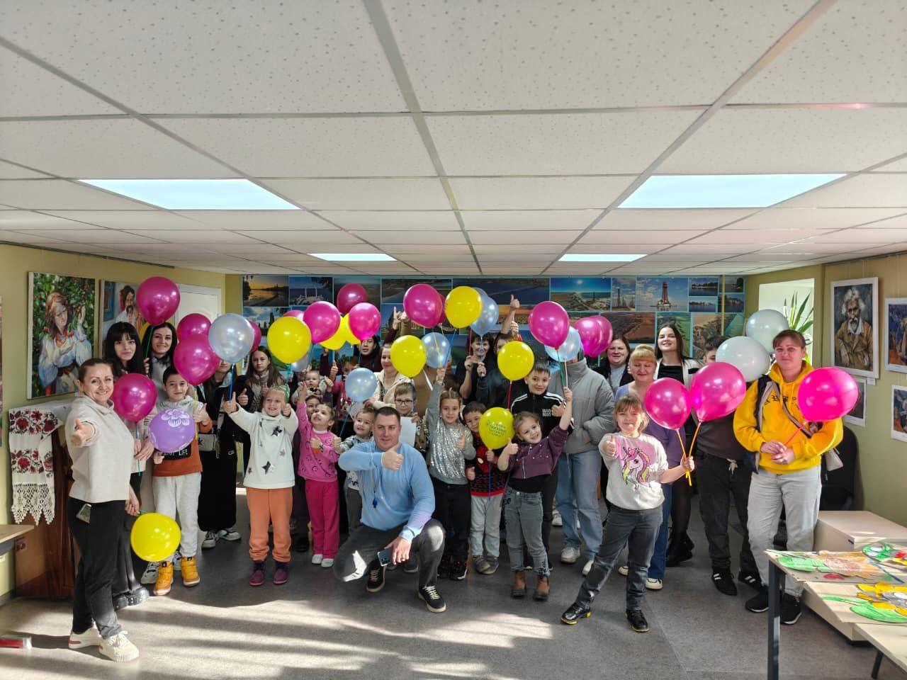 Велика група дорослих і дітей у світлій кімнаті, оточені рожевими, жовтими та сірими повітряними кульками. Більшість людей посміхаються і показують великі пальці вгору. На стінах висять картини.