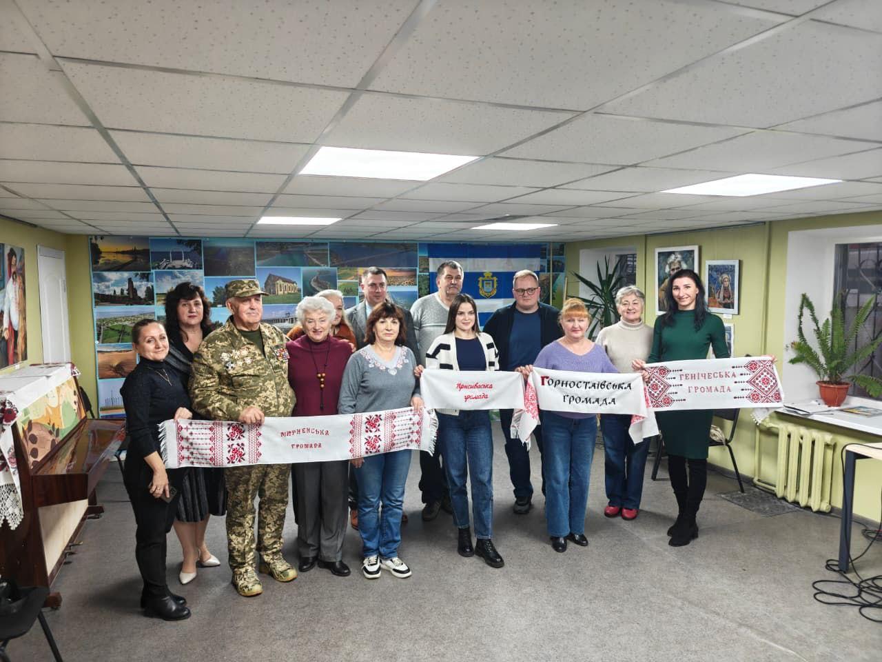 13 осіб, серед яких військовий у формі, стоять у кімнаті, тримаючи вишиті рушники з написами: Виноградівська громада, Краснопільська громада, Горностаївська громада та Генічеська громада.