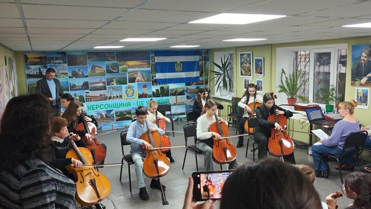 Концертний виступ, де близько десяти дітей грають на віолончелях. На стіні за ними висить великий колаж із пейзажами та гаслом: Херсонщина – це Україна поруч із прапором.