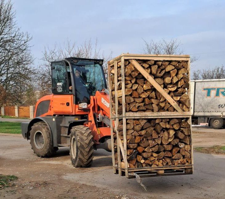 Помаранчевий колісний навантажувач переміщує великий дерев'яний ящик, наповнений дровами, на відкритій дорозі.