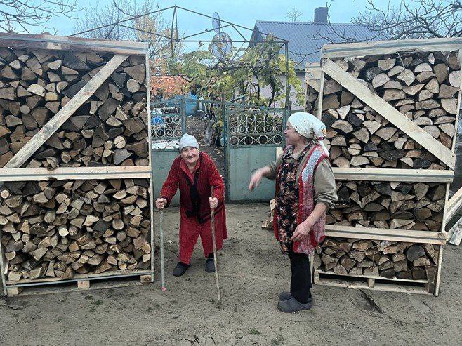 Дві жінки літнього віку, стоять між двома великими дерев'яними ящиками, наповненими дровами. Одна жінка одягнена в червоне і спирається на дві палиці, інша в хустці та фартуху.