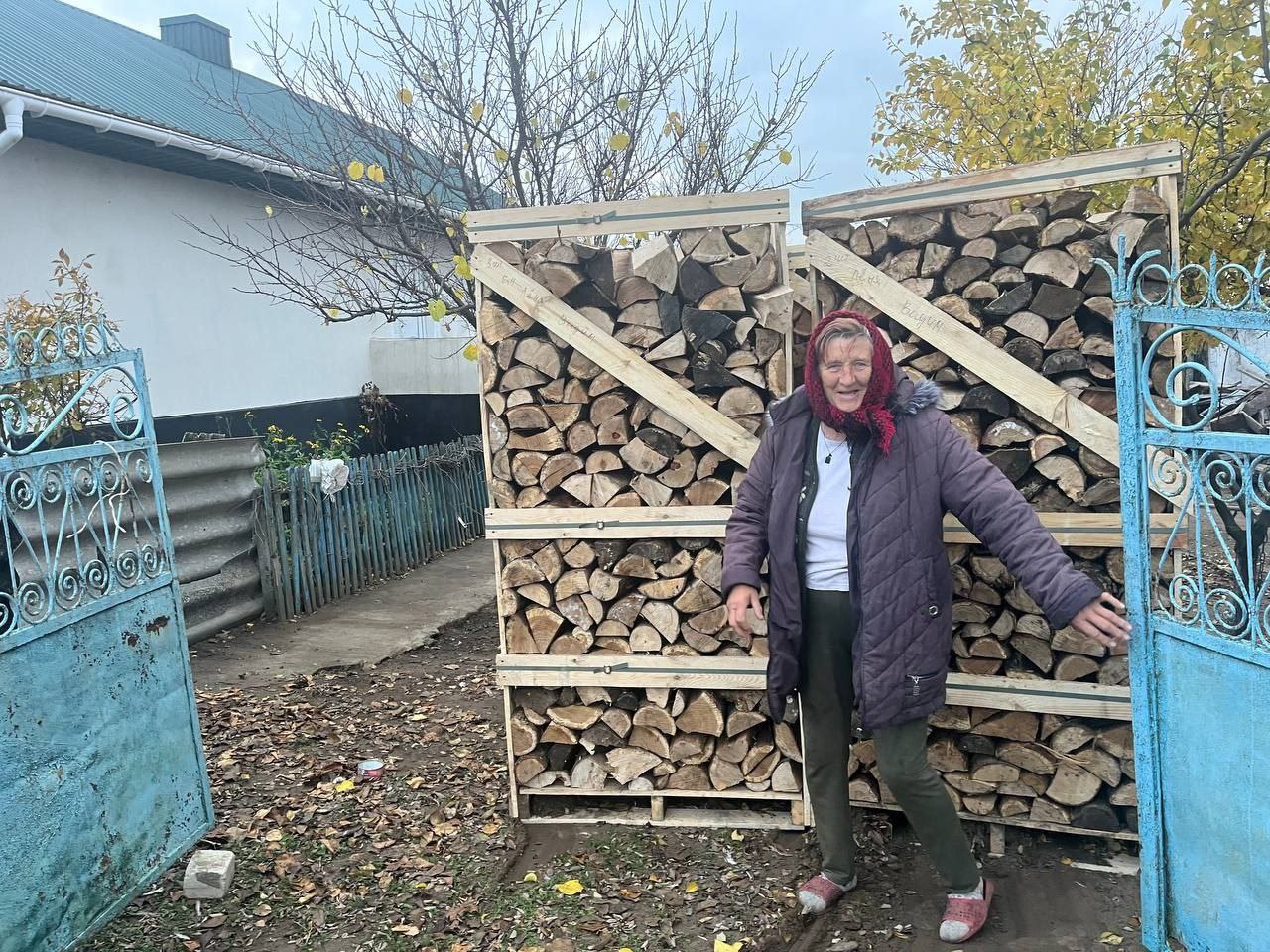 Літня жінка у фіолетовій куртці та червоній хустці стоїть біля відкритих синіх воріт, поруч із двома високими дерев'яними ящиками, наповненими дровами.