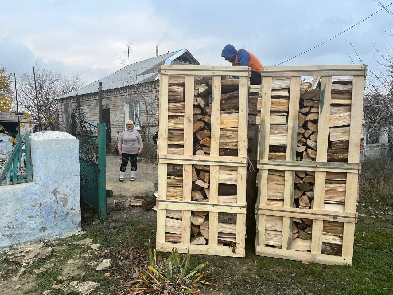 Два великі дерев'яні ящики, наповнені дровами, стоять на ґрунтовій ділянці. Чоловік у помаранчевій куртці знаходиться нагорі правого ящика, а жінка у світлій кофті стоїть ліворуч біля зелених воріт.