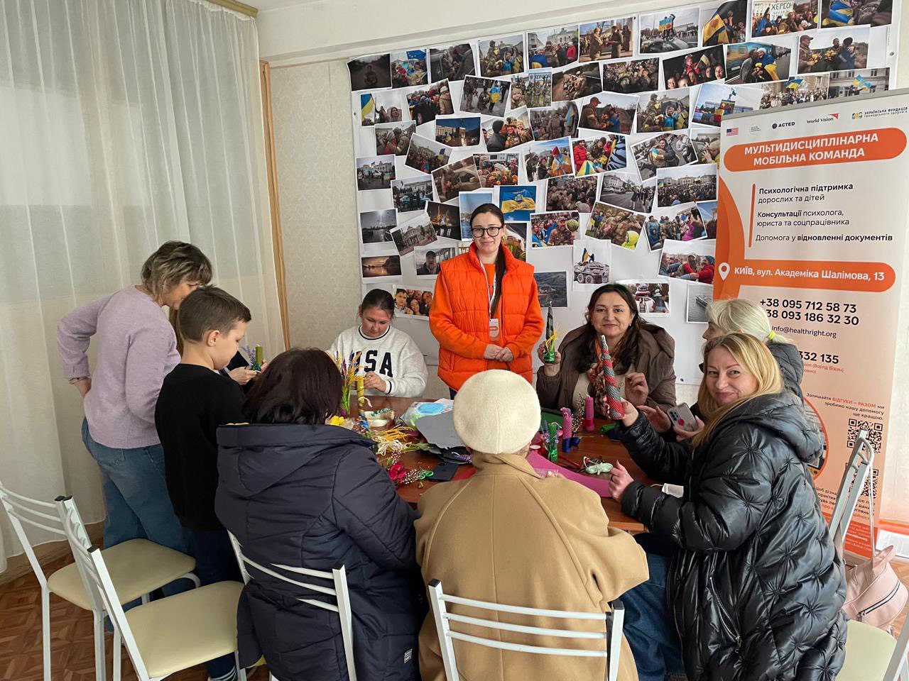 Група людей, включаючи дорослих та дітей, зібралася навколо столу для рукоділля, тоді як жінка в помаранчевій куртці стоїть поруч. На стіні позаду них розміщено великий колаж із фотографій та інформаційний банер про Мультидисциплінарну мобільну команду з контактною інформацією та послугами.