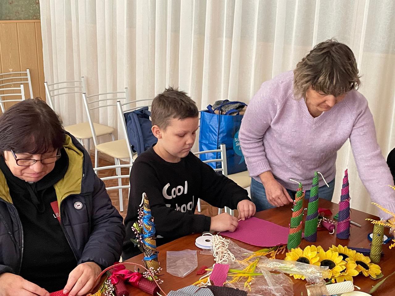 Крупний план трьох людей, які беруть участь у майстер-класі з рукоділля, виготовляючи свічки або декоративні елементи. Хлопчик у центрі в чорному світшоті зосереджено працює з рожевим матеріалом, а дві жінки поруч з ним займаються своїми виробами, серед яких є вже готові спіральні елементи та декоративні соняшники.