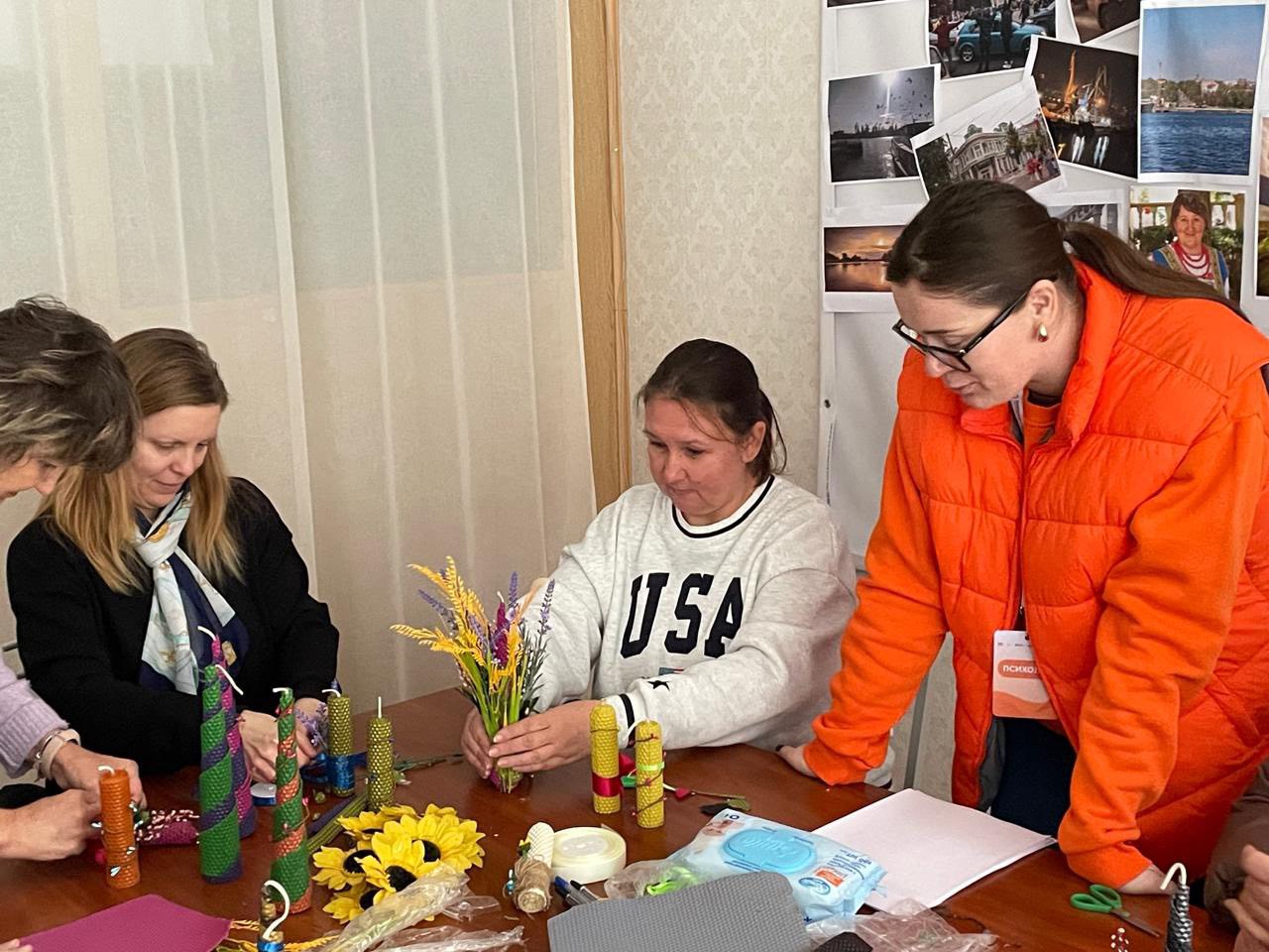 Крупний план чотирьох жінок, які сидять або стоять навколо столу під час майстер-класу з декорування, ймовірно, свічок. Жінка в центрі, у світшоті з написом: USA, тримає букет з жовтих і фіолетових сухоцвітів. Стіл заставлений матеріалами для рукоділля та готовими спіральними свічками.