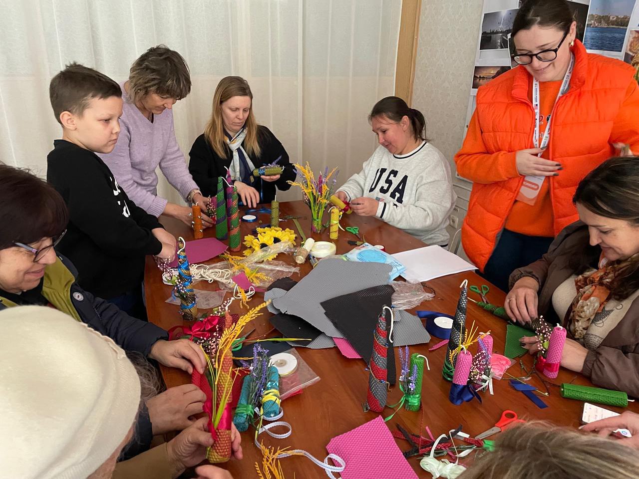 Група з восьми дорослих і однієї дитини зібралася навколо дерев'яного столу, зосереджено займаючись рукоділлям, декоруванням або виготовленням яскравих спіральних свічок із вощини. На столі багато матеріалів, включаючи стрічки, сухоцвіти, листи воску та ножиці.