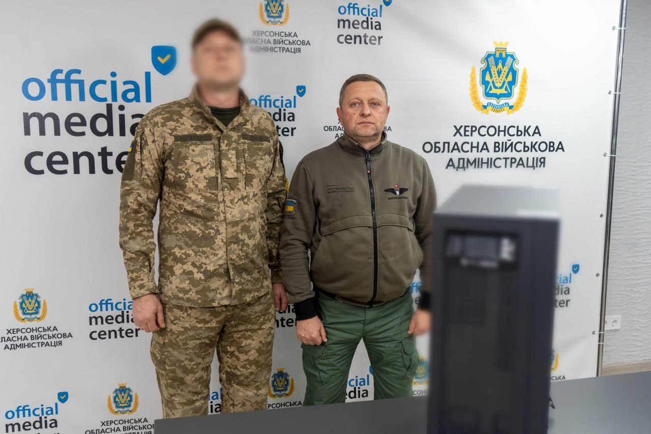 Двоє чоловіків у військовій формі стоять перед фоном з логотипом Херсонської обласної військової адміністрації. Праворуч — чорний пристрій, схожий на джерело безперебійного живлення або інше обладнання.