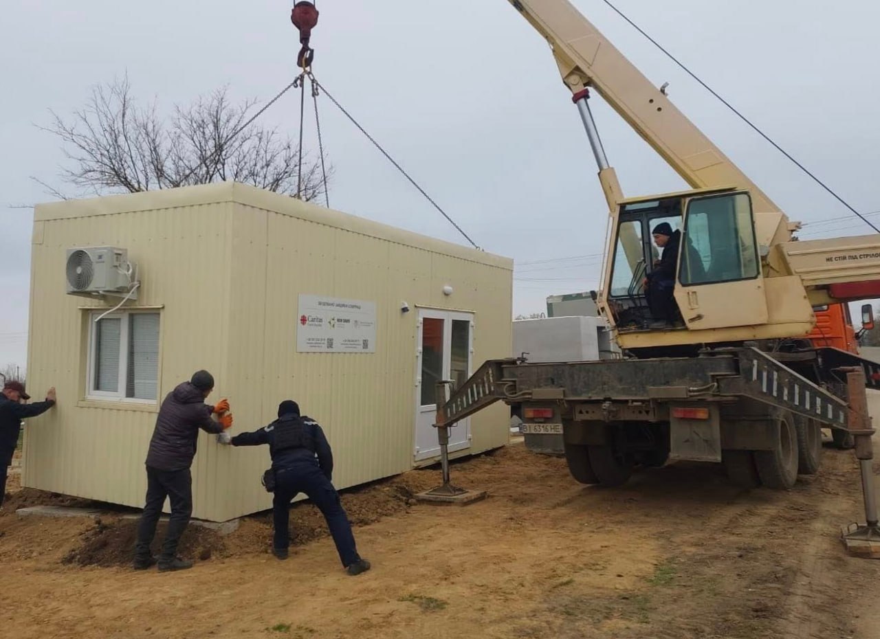 Мобільний кран піднімає і встановлює бежеву модульну будівлю (імовірно, медичний пункт або центр допомоги) на ґрунтовій ділянці. Кілька робітників допомагають спрямувати конструкцію. На стіні будівлі видно логотипи благодійних організацій, зокрема Caritas.