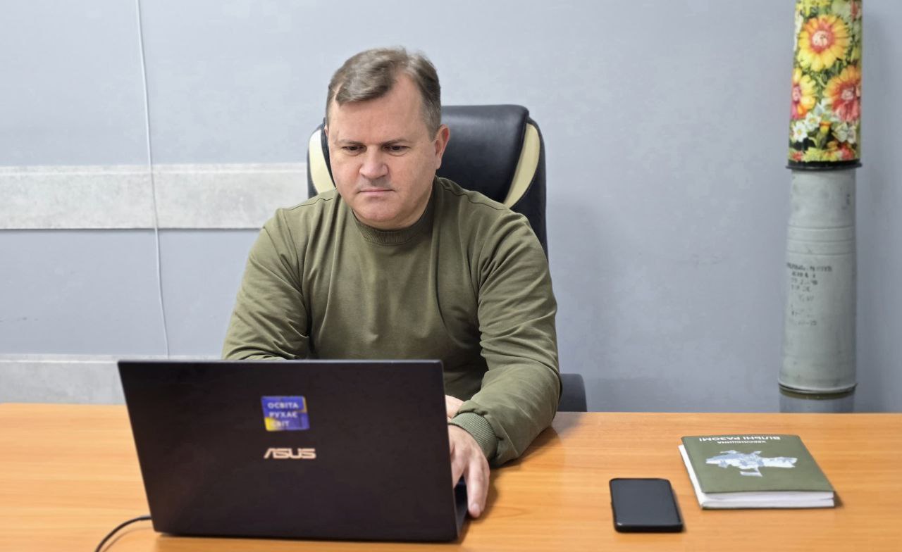 Чоловік середніх років у військовій формі або светрі кольору хакі сидить за робочим столом, зосереджено дивлячись на відкритий ноутбук ASUS. На столі також лежить смартфон та блокнот або книга з написом: Вічні вартості. На задньому плані видно два артилерійські снаряди, один з яких прикрашений квітковим орнаментом.