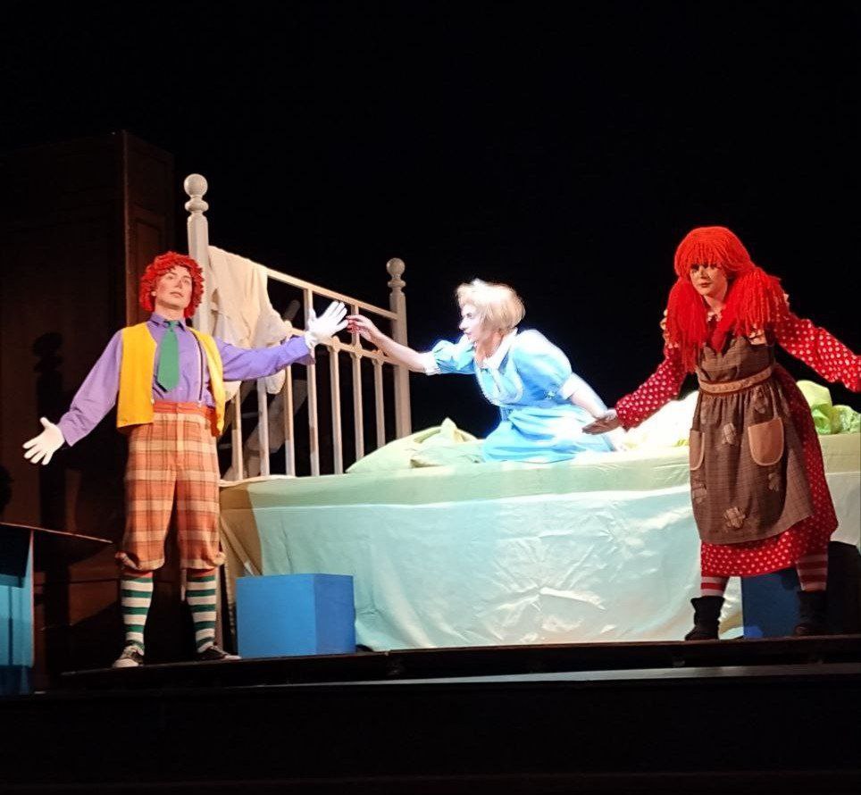 Сцена з театральної вистави, де троє акторів стоять або сидять на сцені. Жінка зі світлим волоссям у блакитній сукні сидить на великому ліжку. Зліва стоїть актор у костюмі з рудим волоссям, жовтим жилетом та смугастими панчохами, а справа – акторка у довгій сукні з рудою кучерявою перукою. Усі троє витягують руки, ніби взаємодіючи один з одним.