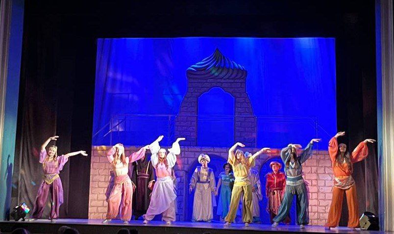 Сцена з театральної вистави на східну тематику, ймовірно, мюзиклу, де група акторів у яскравих, кольорових східних костюмах виконує танцювальний номер. Вони стоять у ряду з піднятими та витягнутими руками, на тлі яскраво-синього задника та декорацій, що імітують кам'яну стіну з аркою та куполом.