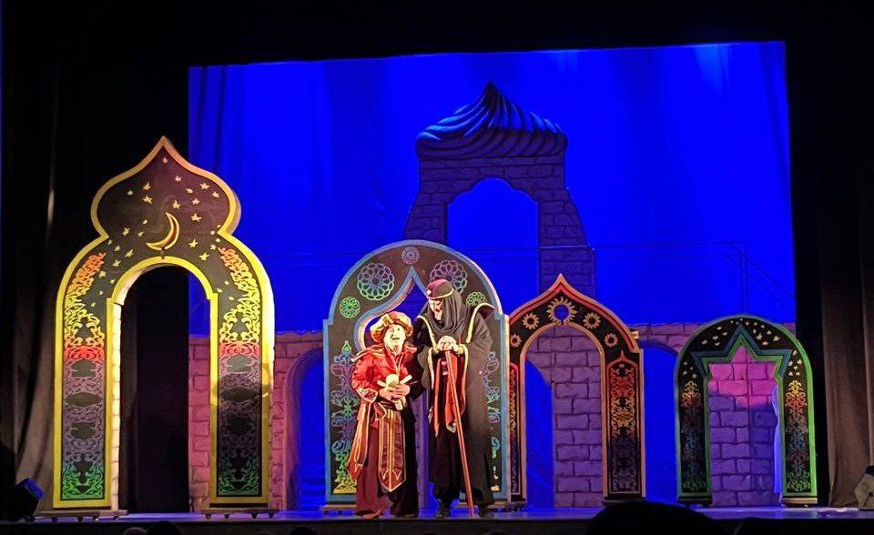 Сцена з театральної вистави на східну тематику. На сцені стоять двоє акторів у багатих східних костюмах (один у червоному, інший у темному), на тлі яскраво-синього задника та декорацій, що зображують стилізовані арки та куполи з ісламськими візерунками.