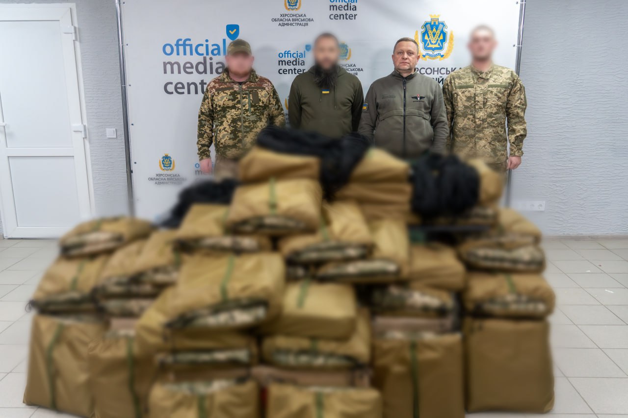 Четверо чоловіків, двоє з яких у військовій формі, стоять на фоні банера Official Media Center Херсонської ОВА позаду великої купи упакованого спорядження. Обличчя трьох чоловіків приховані.