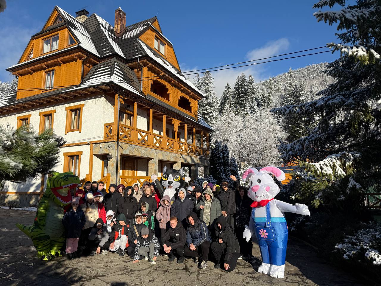 Велика група дітей у зимовому одязі стоїть на вулиці перед багатоповерховим будинком у карпатському стилі. Поруч із дітьми стоять аніматори в костюмах динозавра, кота та кролика. Погода сонячна, навколо лежить сніг.