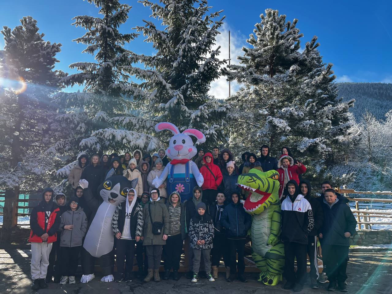 Велика група дітей та підлітків позує разом із ростовими ляльками (сірим котом, білим зайцем та зеленим динозавром) на фоні високих засніжених ялинок у сонячний день.