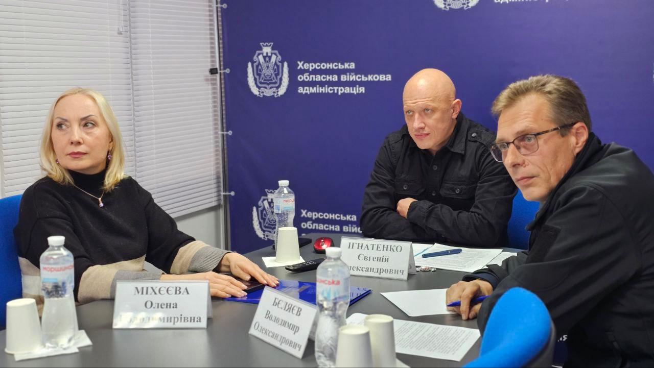 Жінка та двоє чоловіків сидять за столом із документами та іменними табличками на фоні банера Херсонської обласної військової адміністрації.