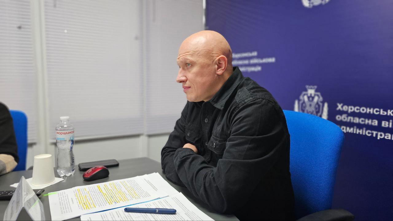 Лисий чоловік у темному одязі сидить за столом із документами на фоні банера Херсонської обласної військової адміністрації.