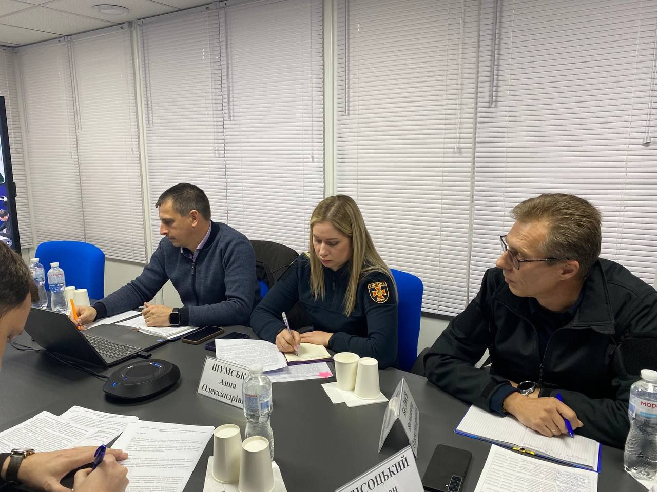 Кілька людей сидять за довгим чорним столом у світлій кімнаті для нарад, роблячи записи на паперах. Зліва направо: чоловік у синьому светрі працює на ноутбуці; жінка зі світлим волоссям у темно-синій формі з емблемою сидить, записуючи щось у блокнот, праворуч сидить чоловік в окулярах і чорній куртці. На столі лежать документи, пляшки з водою та одноразові стаканчики. На задньому плані — білі горизонтальні жалюзі на вікнах.