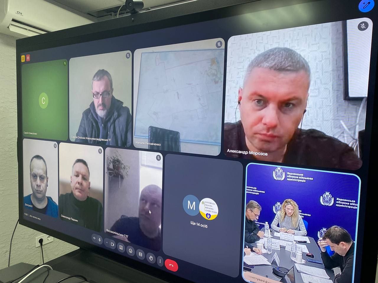 Крупний план великого монітора, на якому відображено відеоконференцію з багатьма учасниками у сітковому режимі. У нижньому правому куті екрана розташовано велике відео, яке показує групу людей, що сидять за столом, на фоні видно емблему та напис: Херсонська обласна військова адміністрація.