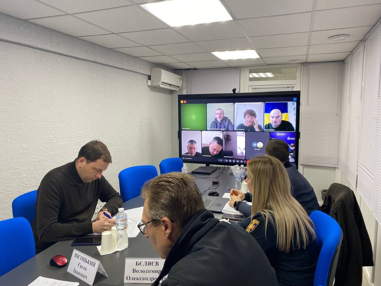 Крупне зображення чоловіка в чорному светрі, який сидить за довгим столом у світлому офісі, схилившись над паперами. Поруч з ним сидять ще двоє людей, а на великому настінному моніторі проходить відеоконференція з чотирма учасниками.