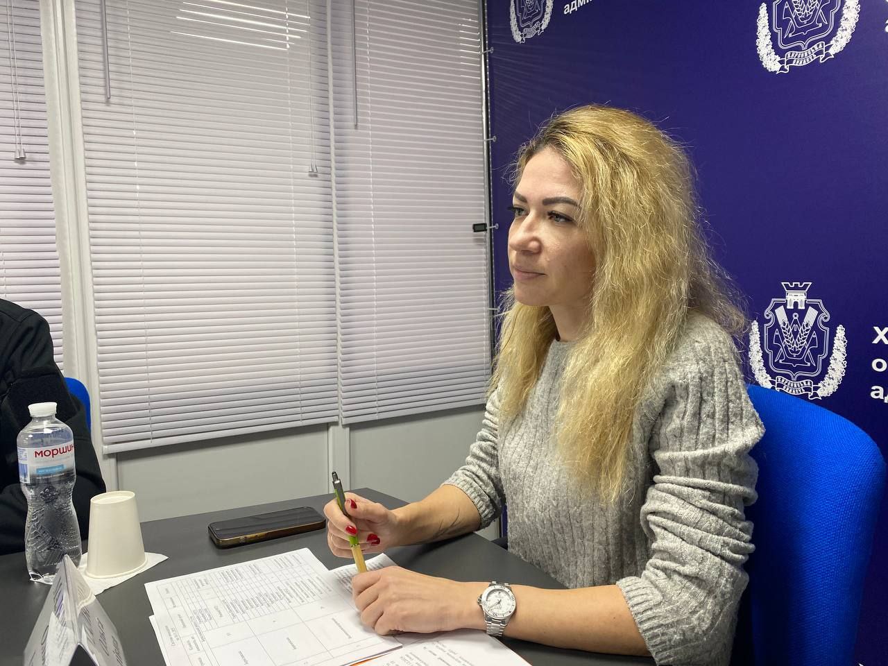 Жінка зі світлим волоссям у сірому светрі сидить за столом з паперами та олівцем, уважно слухаючи.