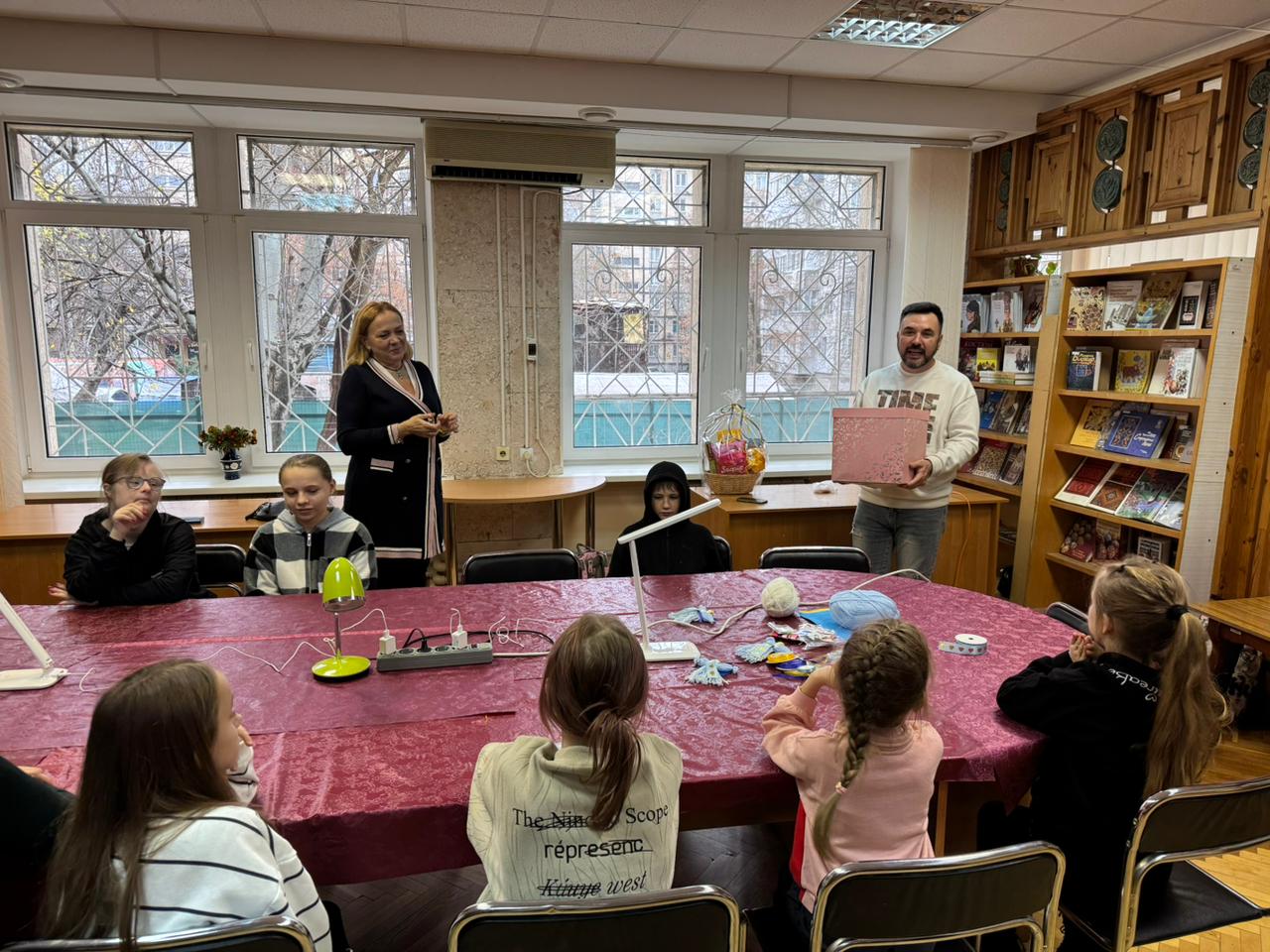 Група дітей сидить за довгим столом, слухаючи двох дорослих, що стоять поруч із великими вікнами та книжковими шафами.