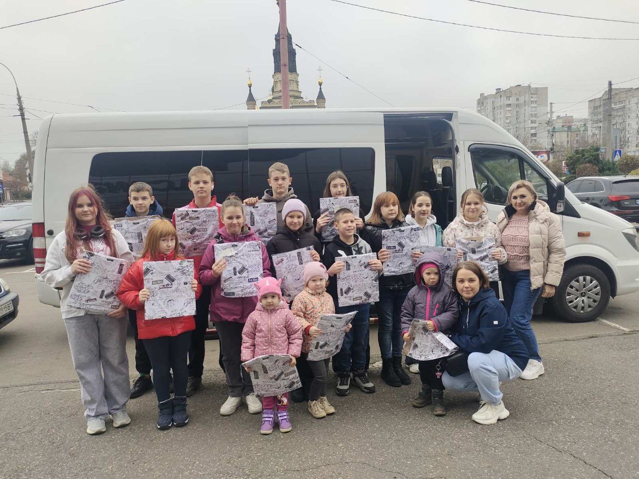 Група з приблизно десяти дітей та кількох дорослих позує для фотографії, стоячи на тлі великого червоно-коричневого рекламного банера Одеського державного цирку з написами: ЦІКАВІ ЦИФРИ та інформацією про місткість залу.