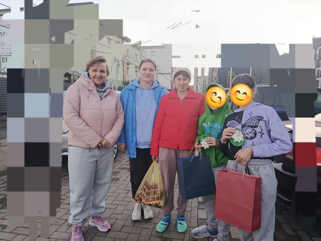 Групове фото на вулиці, де чотири жінки та двоє дітей стоять, тримаючи пакети та подарунки, ймовірно, після отримання допомоги.