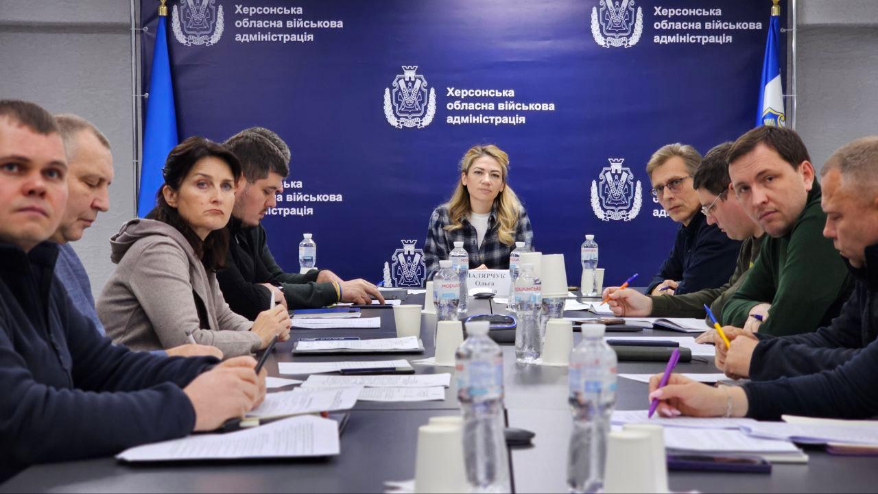 Нарада або зустріч за круглим столом, на якій присутні близько десяти людей. Жінка в центрі — голова, сидить навпроти учасників. На задньому плані — синій банер з логотипом і написом: Херсонська обласна військова адміністрація.