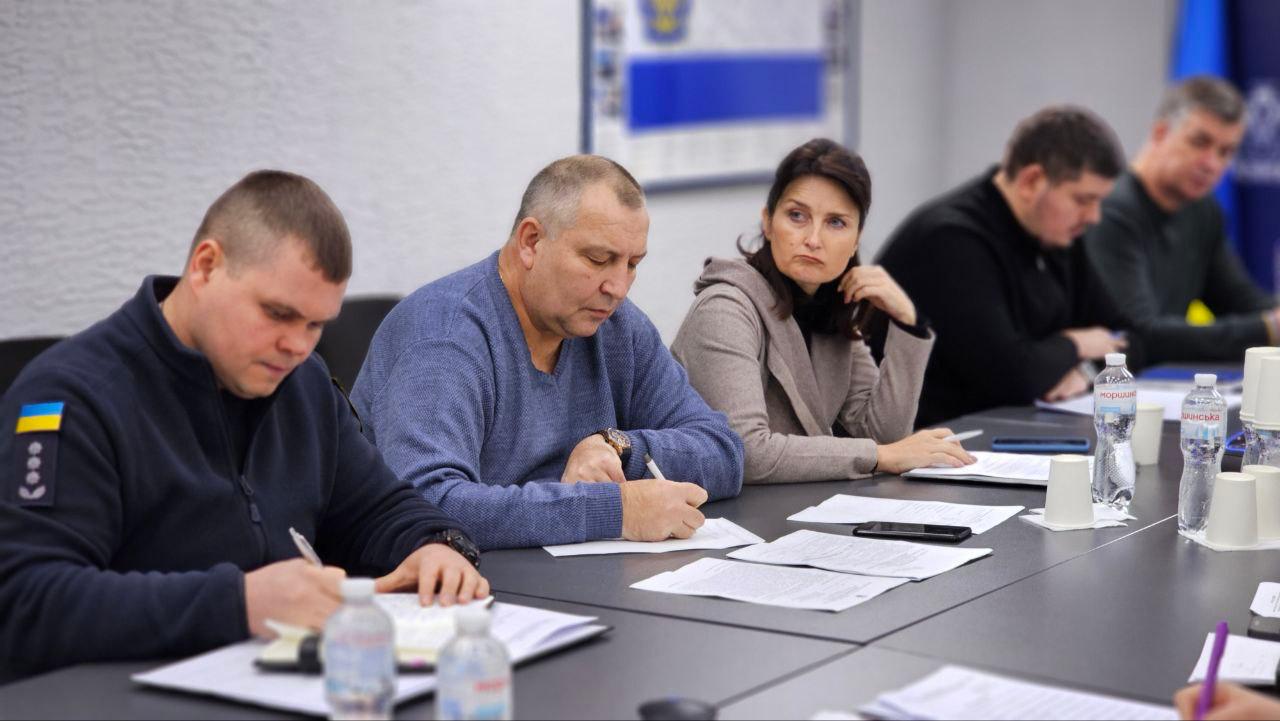 Крупний план частини наради, де троє чоловіків, включно з одним у синій кофті та одним у темній формі з українським прапором на рукаві, зосереджено пишуть на паперах, що лежать на столі. Поруч сидить жінка. На столі видно пляшки з водою.