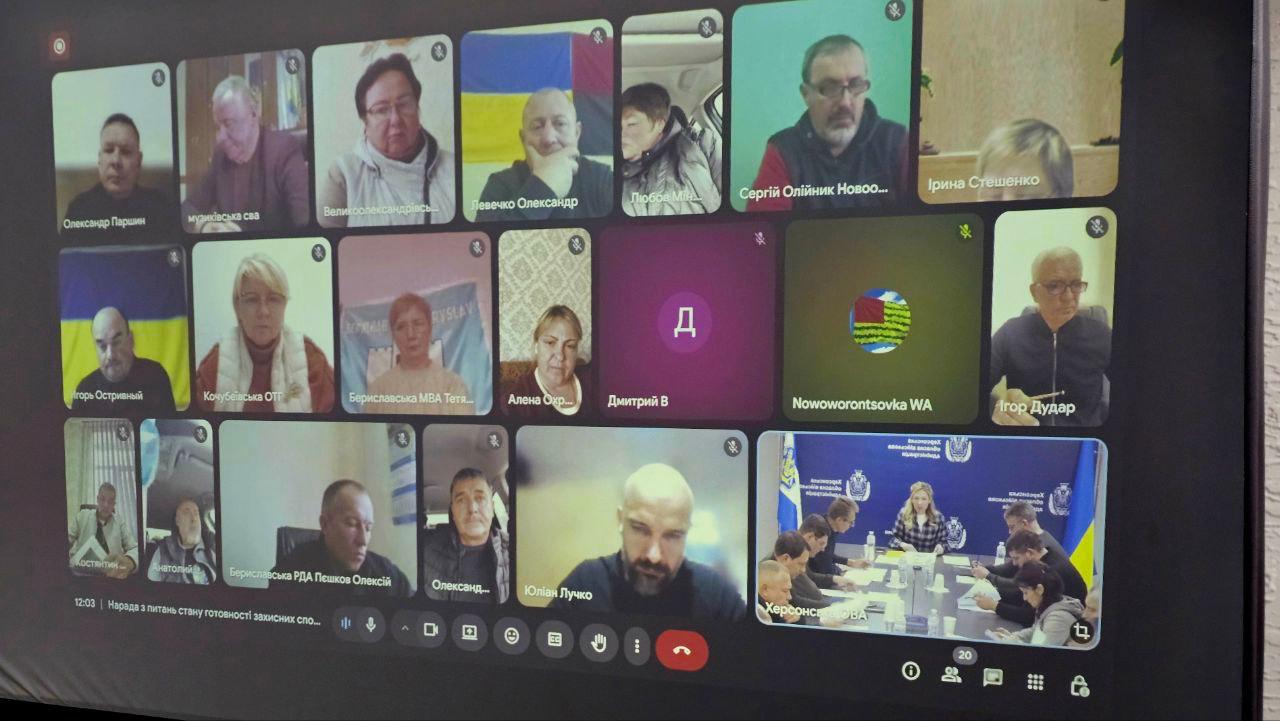 Екран монітора з відображенням відеоконференції (онлайн-наради) у форматі сітки. На плитках видно обличчя багатьох учасників, деякі з іменами або назвами установ. В одному з вікон видно учасників, які сидять за столом, підписаних як: Херсонська ОВА.